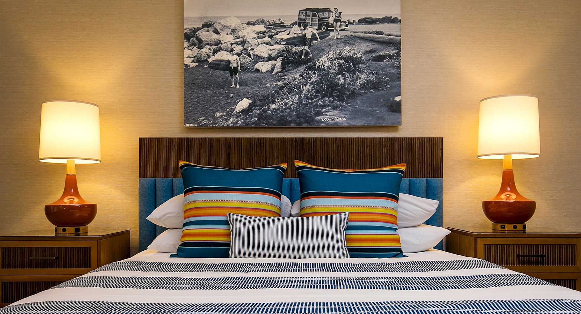 A cozy hotel bedroom with a striped bed, colorful throw pillows, two matching lamps, and a black-and-white wall art above the headboard.