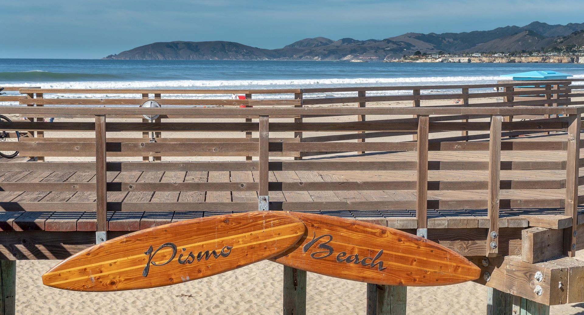 Wooden surfboards hanging on a pier, a sandy beach, blue ocean, and a boardwalk with hills in the distance. Beach vibes at its calm best.