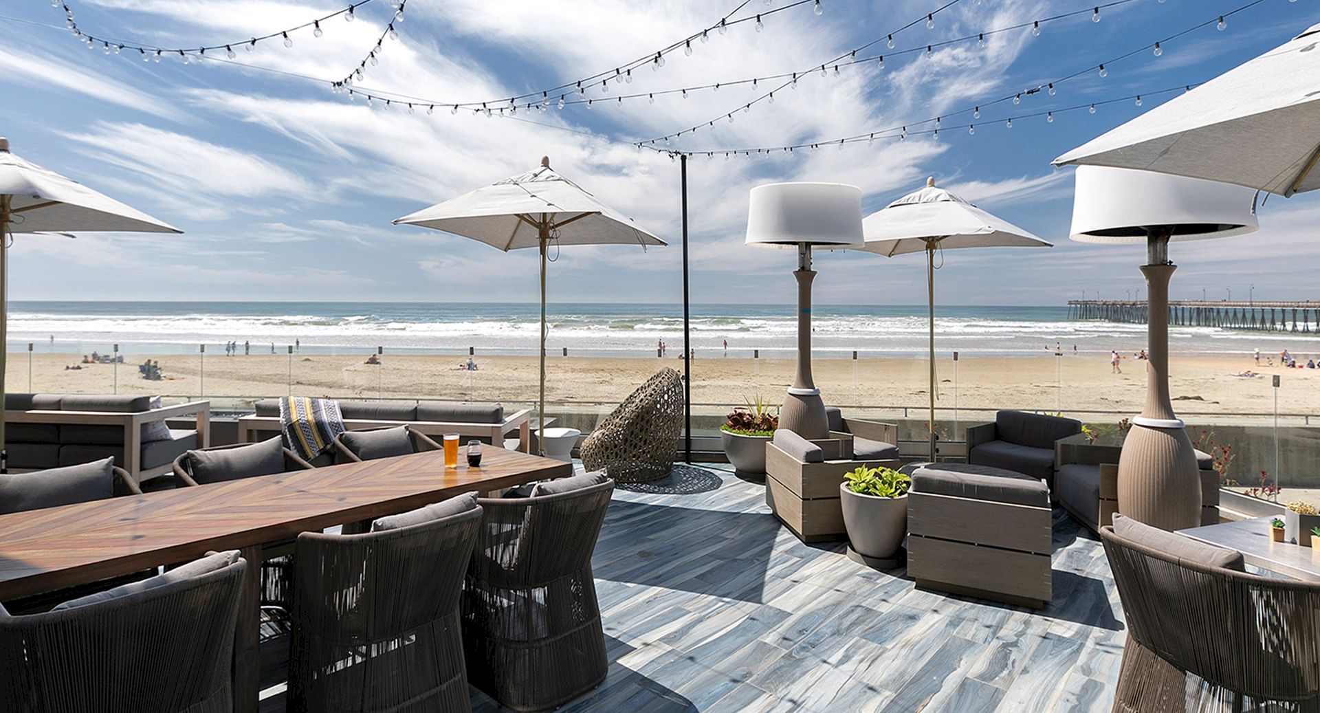Coastal beachside lounge with wicker seating, wooden tables, umbrellas, string lights, and planters, facing the sunny shore and ocean. ending.