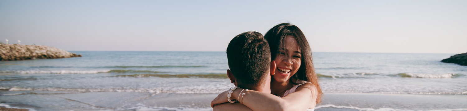 Two people share a joyful hug on a sunny beach, the ocean waves rolling in behind them as they smile warmly.