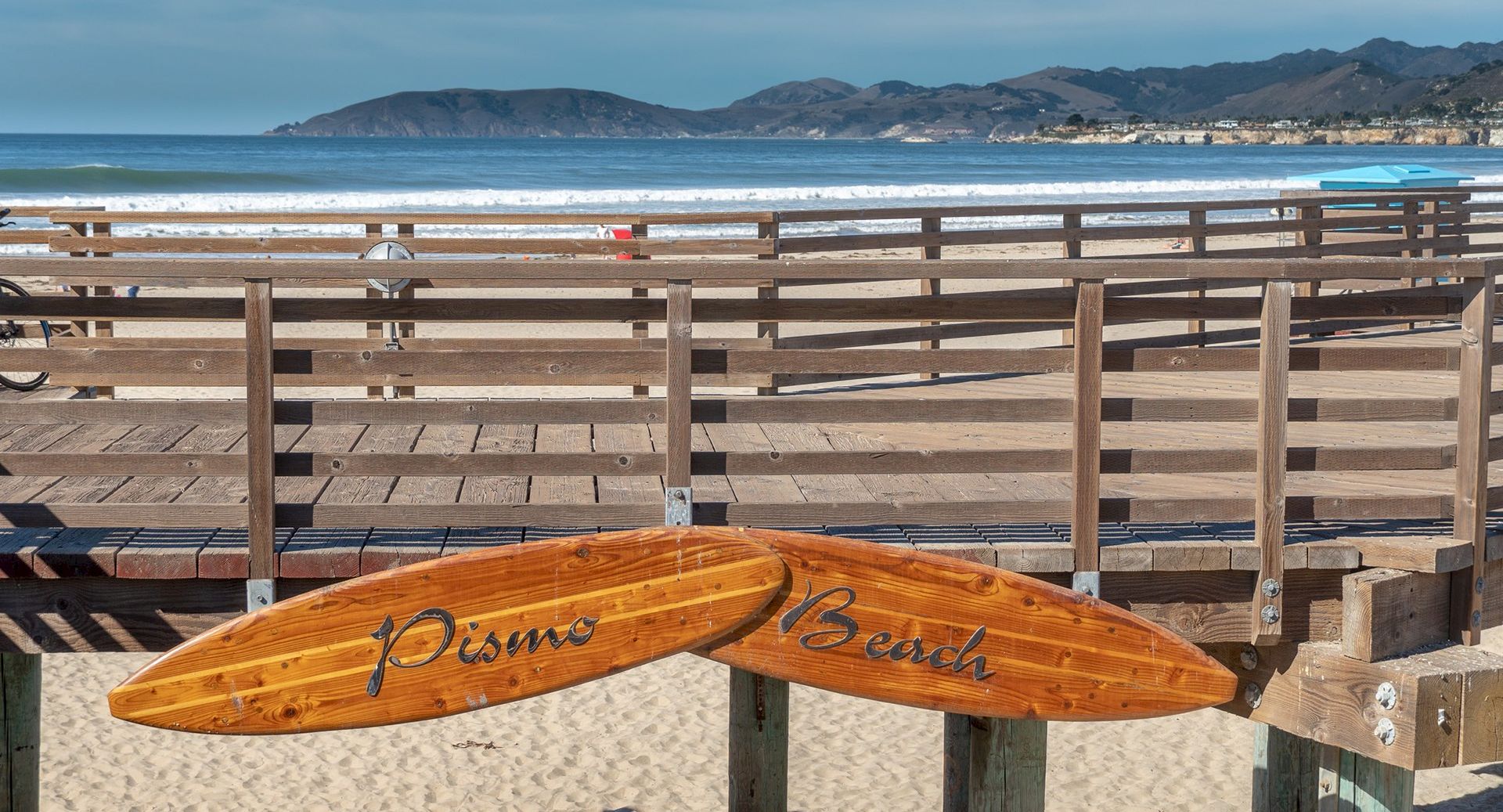A sunny beach scene with a wooden boardwalk over sand, ocean in the background, and two wooden surfboards hanging on the railing.