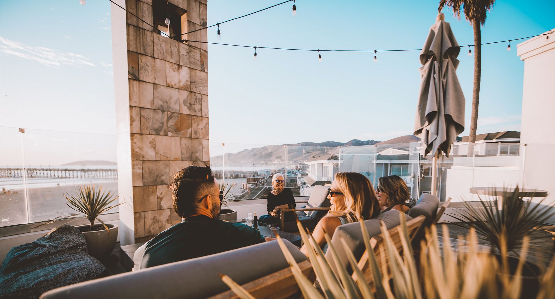 A group of friends relaxing on a patio by the water, chatting around a low table with string lights overhead.