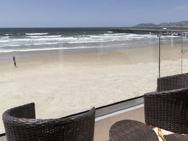 A sunny beach scene with waves, a lone jogger, and a wooden pier in the distance; foreground shows wicker chairs on a patio overlooking the sand.