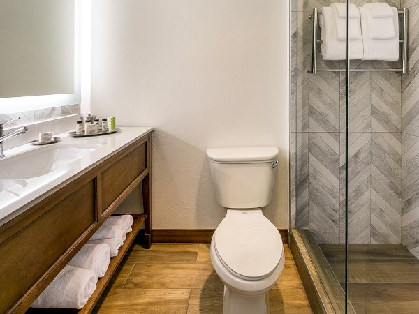 A modern bathroom with a long wooden vanity, double sink, open towel storage, a white toilet, and a glass-enclosed shower with chevron tile walls.