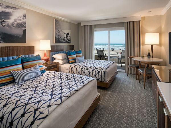 A hotel bedroom with two double beds, blue patterned bedding, seaside-themed photos, lamps, a desk, and a balcony overlooking the ocean, bright and inviting.