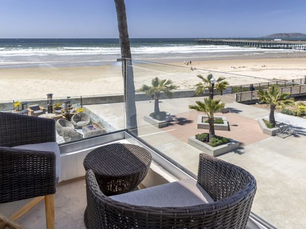 A beach view from a balcony with wicker chairs, a glass railing, palm trees, and a pier extending into the ocean.