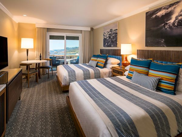 A hotel room with two double beds, striped blue-gray bedding, colorful throw pillows, a flat-screen TV, desk, chairs, lamps, and a window with a view.