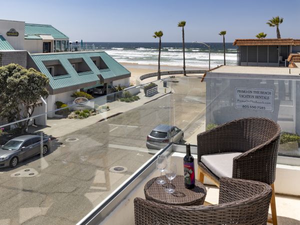 Ocean view from a balcony with wicker chairs, a small table, and a bottle; palm trees line the beach, buildings and cars below.