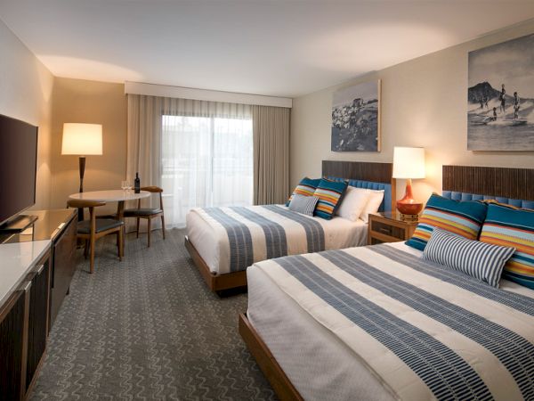 A hotel room with two queen beds, striped blue and gray bedding, bedside lamps, a desk with chair, a TV stand, and large window with sheer curtains.
