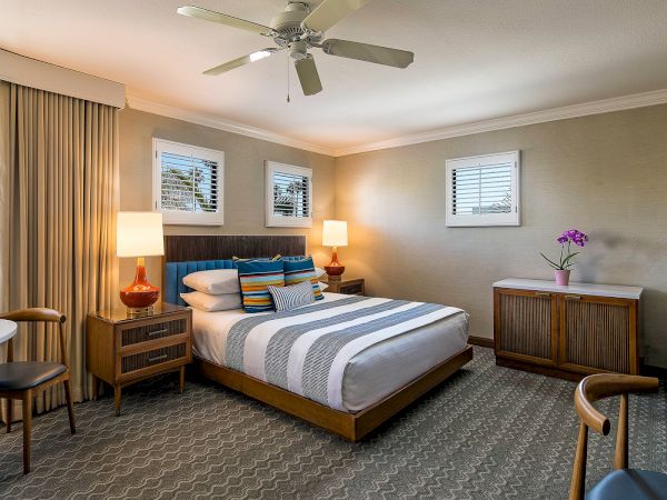 A stylish hotel room with a king bed, striped bedding, two nightstands with lamps, a ceiling fan, a dresser with a vase of flowers, a small table and chairs, and soft natural light from windows in a neutral-toned suite.