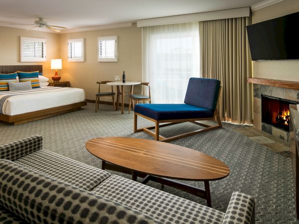 A cozy hotel living area with a bed, sofa, round coffee table, desk, chair, fireplace, wall-mounted TV, and large windows with curtains. End.