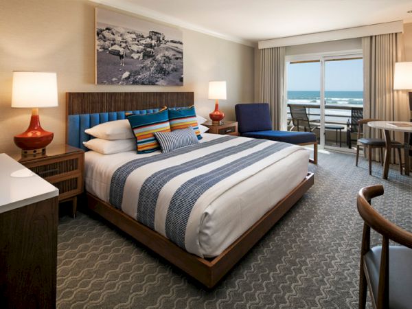 A modern hotel room with a king bed, striped bedding, two nightstands, a seating area, and a balcony overlooking the ocean.