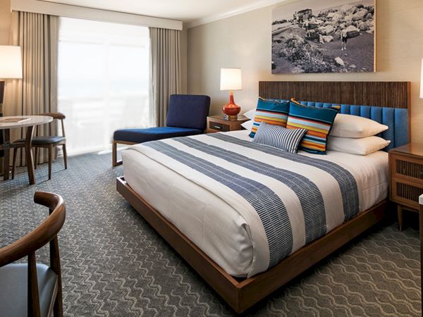 A stylish hotel room with a king bed, blue-accent pillows, bedside lamps, a desk and chair, and a seating area by a large window.