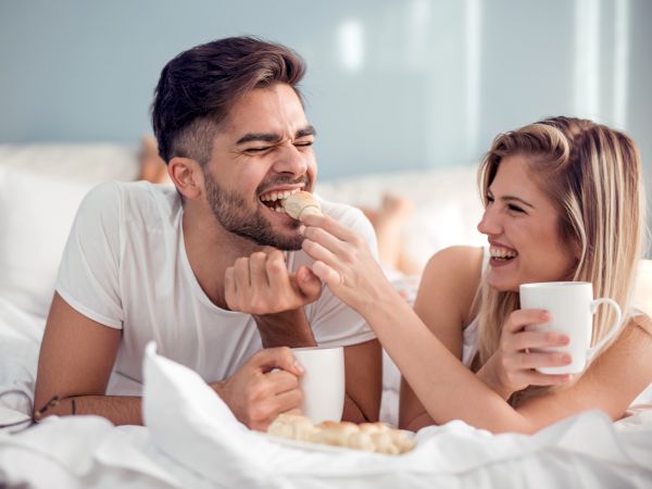 A couple sharing a cozy moment in bed, feeding each other muffins or snacks and sipping hot drinks, smiling and enjoying breakfast together.