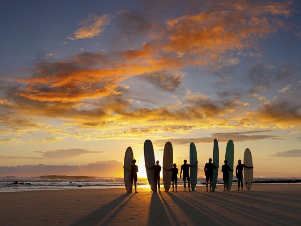 A group of surfers stand on the beach at sunset, surfboards lined up, casting long shadows as orange clouds glow above the ocean.
