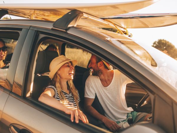 Three friends in a car with surfboards on top, chatting and smiling as they cruise toward the beach at sunset.