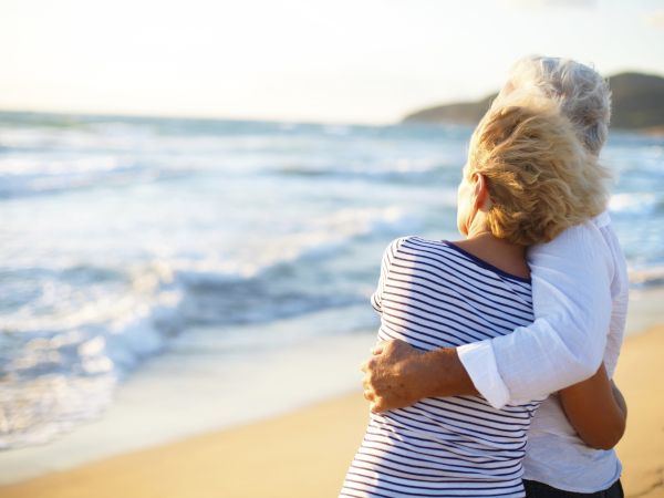 Two people embrace on a sunny beach, waves rolling in, wearing striped shirts and enjoying a warm seaside hug.