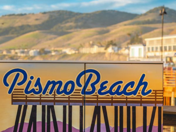 A sign reads &ldquo;Pismo Beach&rdquo; on a beach boardwalk with hills in the background and a sunny coastal scene.