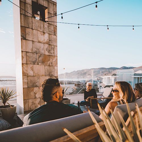 A group of friends relaxing on a patio by the sea, chatting under string lights near the coast.