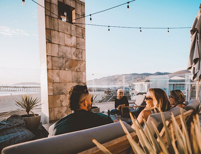 A group of friends relaxing on a patio by the sea, chatting under string lights near the coast.
