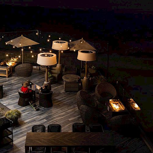 Beachside deck at dusk with string lights, white umbrellas, and a cozy seating area by the water, calm waves beyond.