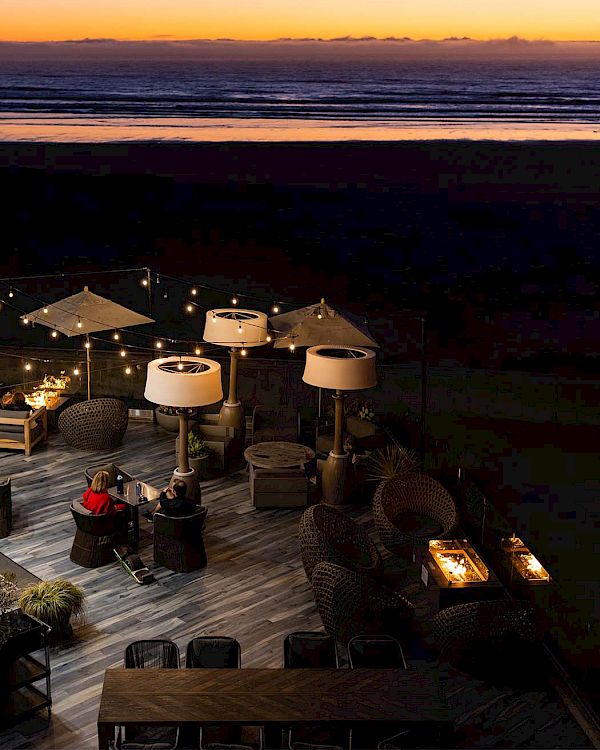 Beachside deck at dusk with string lights, white umbrellas, and a cozy seating area by the water, calm waves beyond.