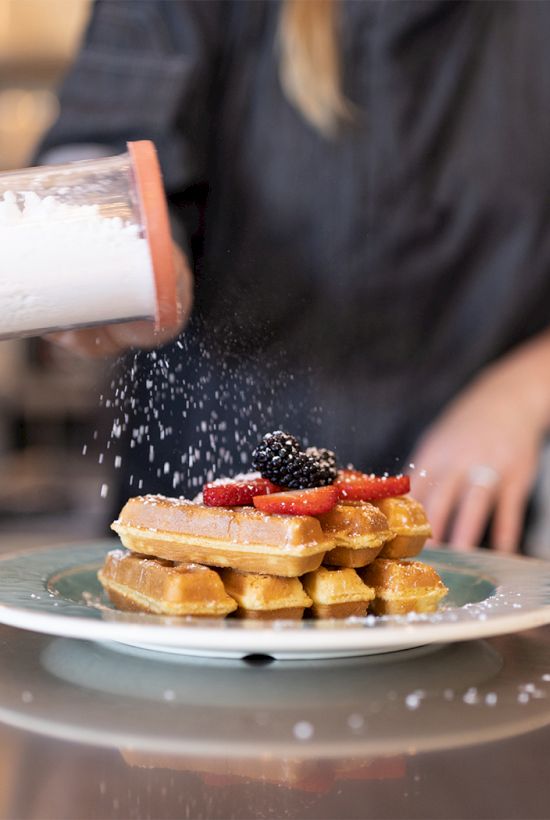 A barista sprinkles powdered sugar on a stacked waffle dessert with strawberry sauce on a plate, while a shaker sprinkles sugar above it.