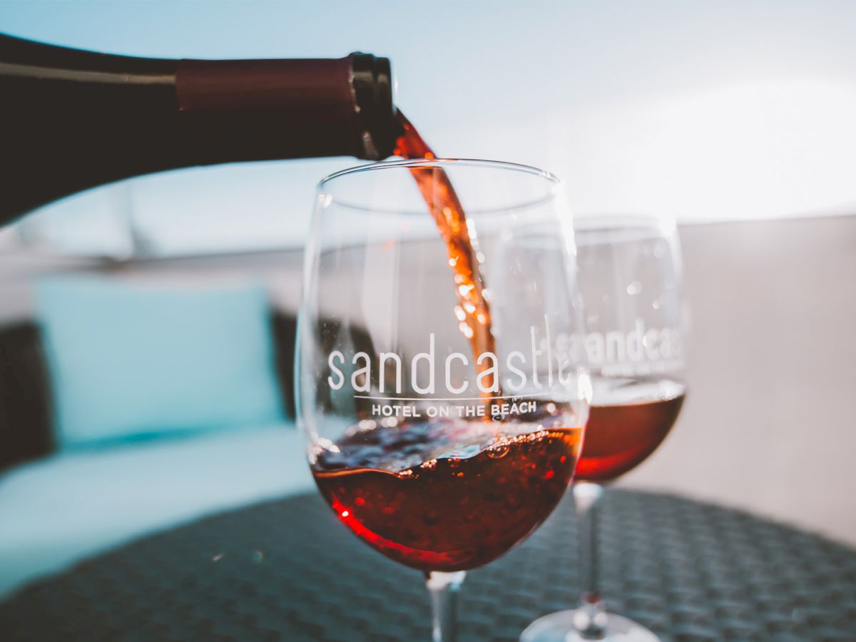 Two glasses of red wine being poured from a bottle, on a seaside or poolside setting with a blurred background.