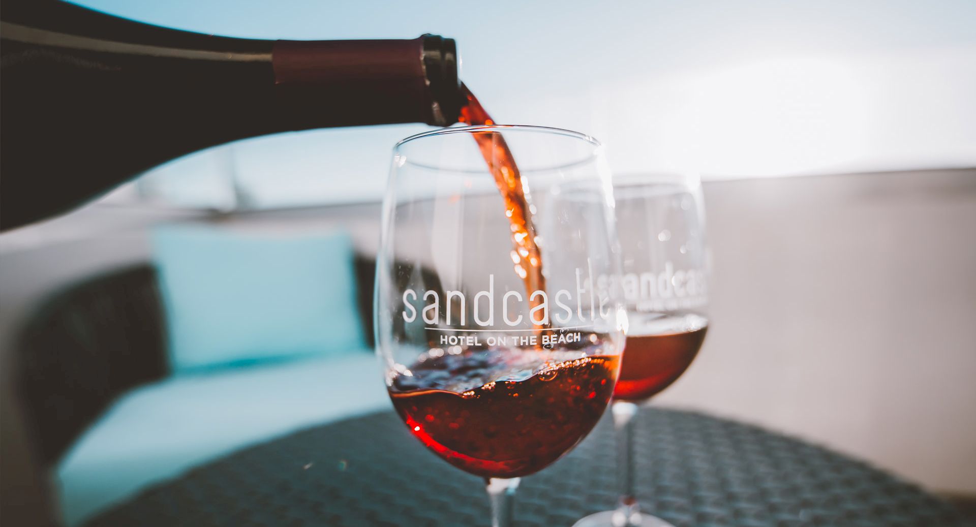 Wine pours into two glasses on a sunny outdoor table at a resort, with &ldquo;sandcas&hellip;.&rdquo; on the glass. Enjoyable, relaxing vibes. ending with a period.