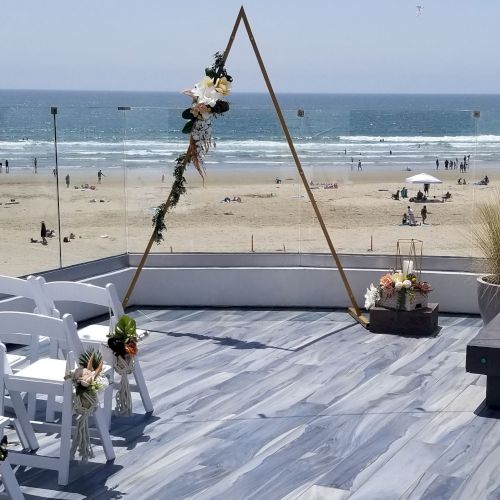 A beach wedding setup with a triangular arch adorned with flowers on a wooden deck, white chairs, and the sea in the background.