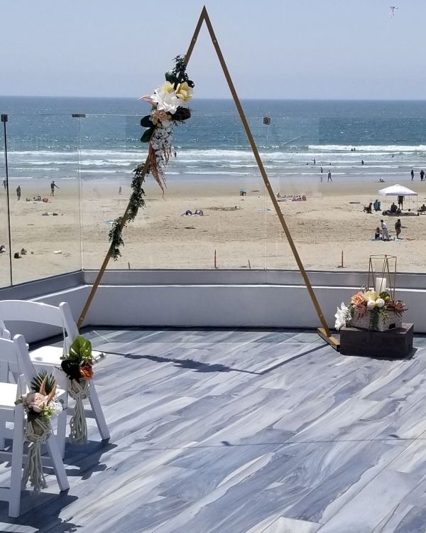 A beach wedding setup with a triangular arch adorned with flowers on a wooden deck, white chairs, and the sea in the background.