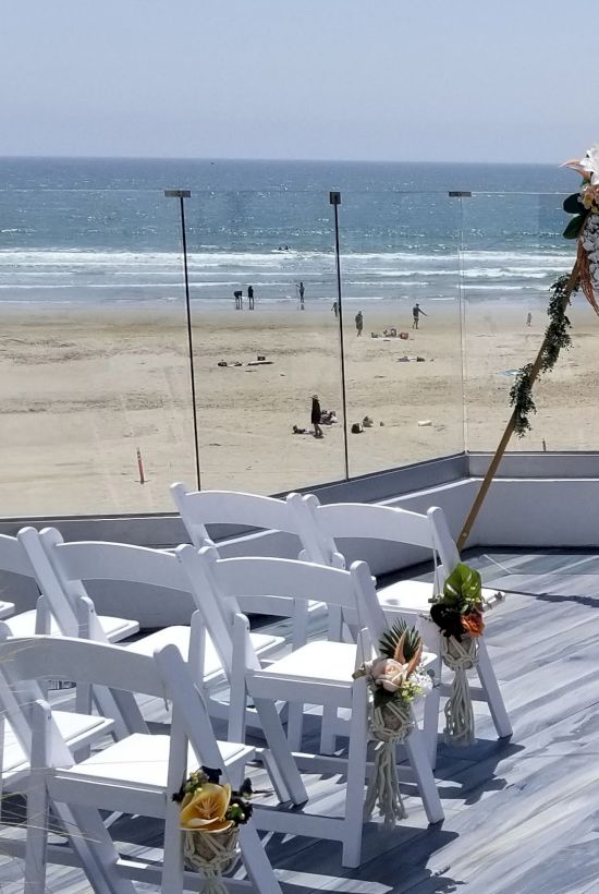 A beach wedding setup with a wooden arch adorned with flowers, white chairs for guests, sandy shore and waves in the background, bright and sunny.