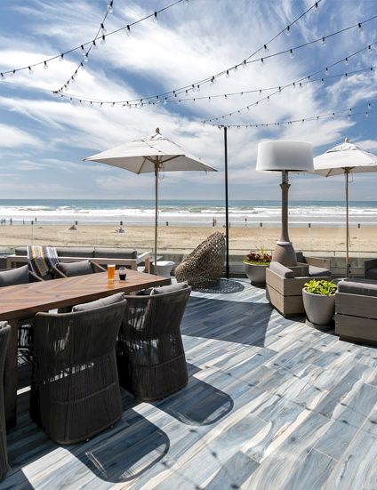 A sunny beachfront outdoor dining area with wicker chairs, long wooden table, umbrellas, string lights, and a sandy shore beyond the deck.