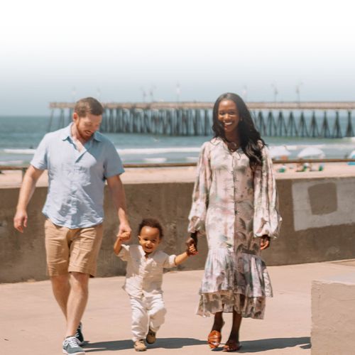 A family strolls on a sunny beach boardwalk: a man, woman, and a small child walk hand-in-hand near the ocean and a pier.