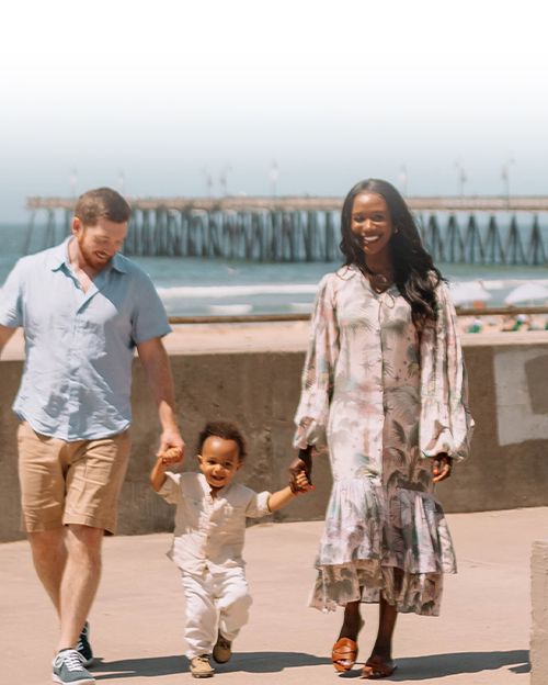 A family strolls on a sunny beach boardwalk: a man, woman, and a small child walk hand-in-hand near the ocean and a pier.