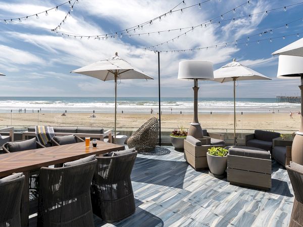 A beachside patio with wicker seating, tables, umbrellas, string lights, and a view of the sandy shore and ocean under a blue sky.