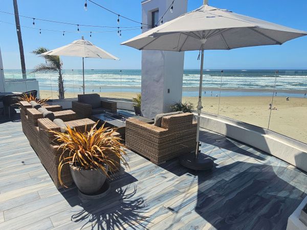 Beachfront patio with wicker seating, glass railing, umbrellas, and string lights; sunny المشهد overlooking waves on a sandy shore.