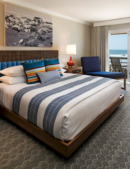 A hotel room with a large bed, blue-striped bedding, two nightstands, lamps, a seating area, and a balcony with a ocean view, all in soft neutrals.