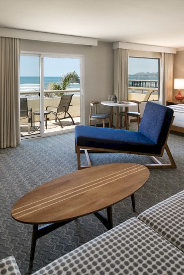 A hotel suite with a cozy seating area, king bed, fireplace, and a balcony overlooking the ocean, airy and stylish with modern furnishings.