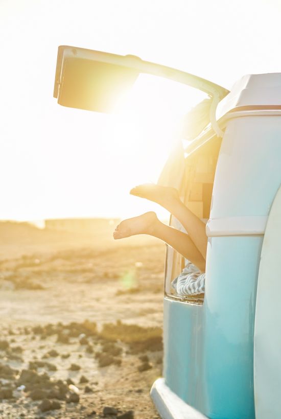 A turquoise retro van on a sunny beach, surfboard strapped to the side, and the sun glowing from behind as waves or dunes roll in.