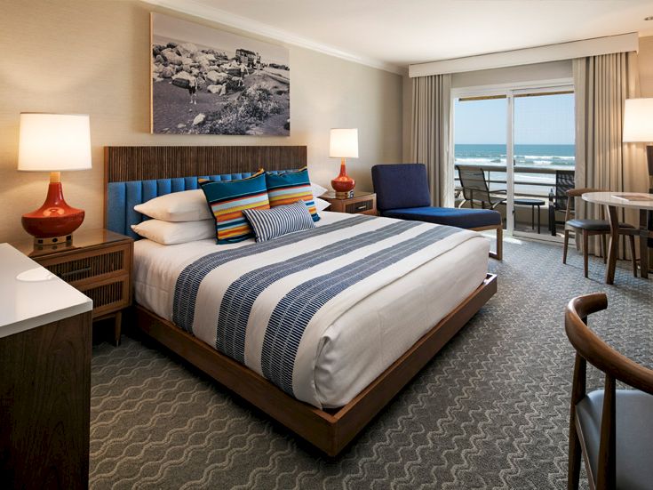 A stylish hotel bedroom with a large bed, striped bedding, two red lamps, a desk, seating area, and a balcony overlooking the ocean.