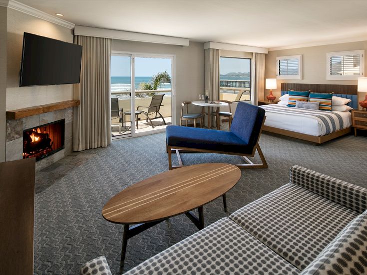 A hotel suite with a fireplace, large windows to an ocean view, a seating area, a round coffee table, and a bed with matching chairs. Top it at 140 characters, always ending the sentence.