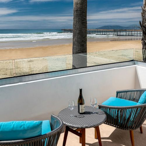 A sunny beach with two lounge chairs, a small table, and an umbrella, calm blue water, and a clear sky in the background.