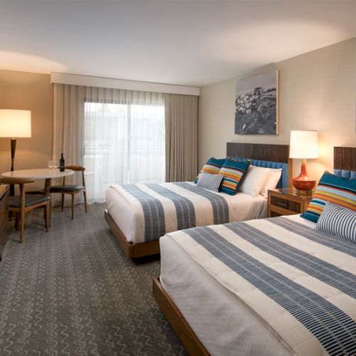 Two double beds in a hotel room with striped blue-gray linens, a desk and chair, a flat-screen TV, a window with sheer curtains, and wall art of winter mountains.