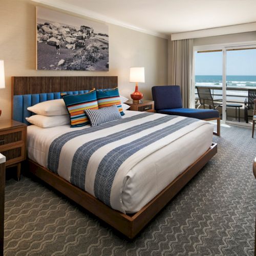 A stylish hotel room with a large bed, ocean view balcony, striped bedding, two lamps, and a seating area, all overlooking the sea.