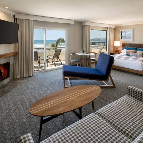 A bright hotel suite with a fireplace, blue-and-gray seating, a desk chair, and a balcony overlooking the ocean; cozy, modern, inviting.