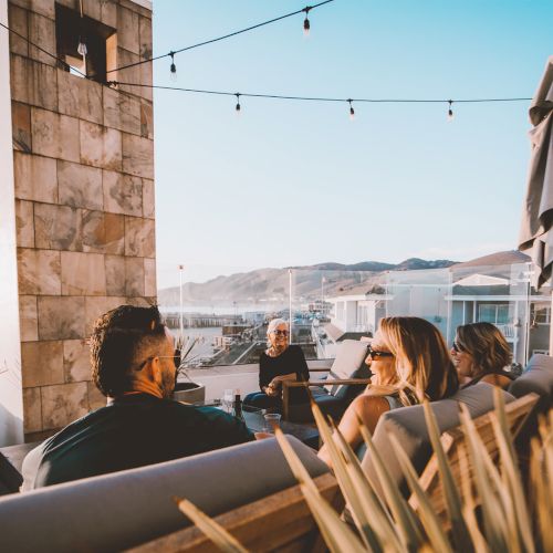 A group of friends sits on outdoor lounge furniture on a sunlit rooftop, chatting by string lights with a view of the sea and hills behind.