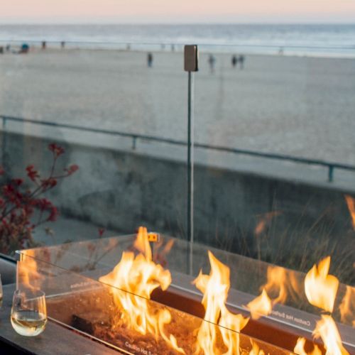 A seaside view with a glass railing, fire pit flames, and two whiskey glasses on a ledge; beach and people in the background at sunset.