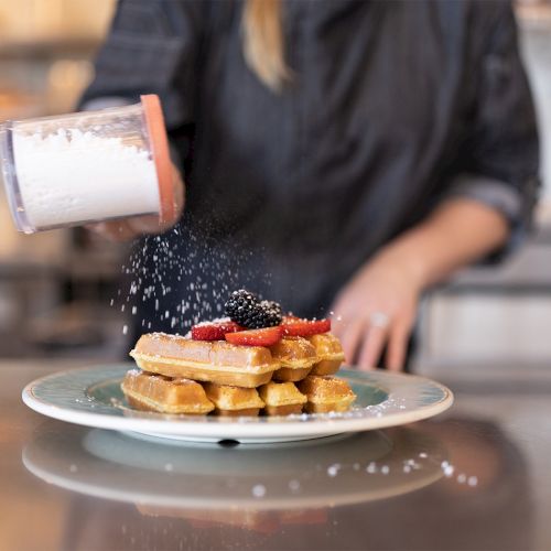A chef sprinkles powdered sugar on a plated waffle stack with berries, as a hand pours sugar from a shaker.