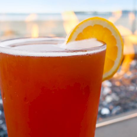 A cocktail with an orange-red hue and a lemon slice on the rim, set against a beachside fire pit backdrop.